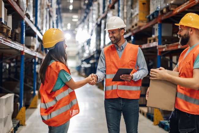 Warehouse workers wearing reflective vests and hard hats shaking hands in a warehouse aisle. One worker is holding a digital tablet