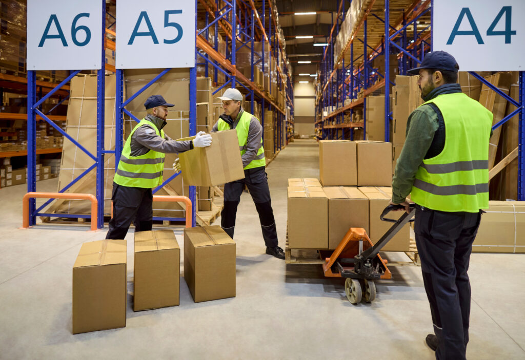 Team of warehouse workers moving boxes with pallet trolley near shelving
