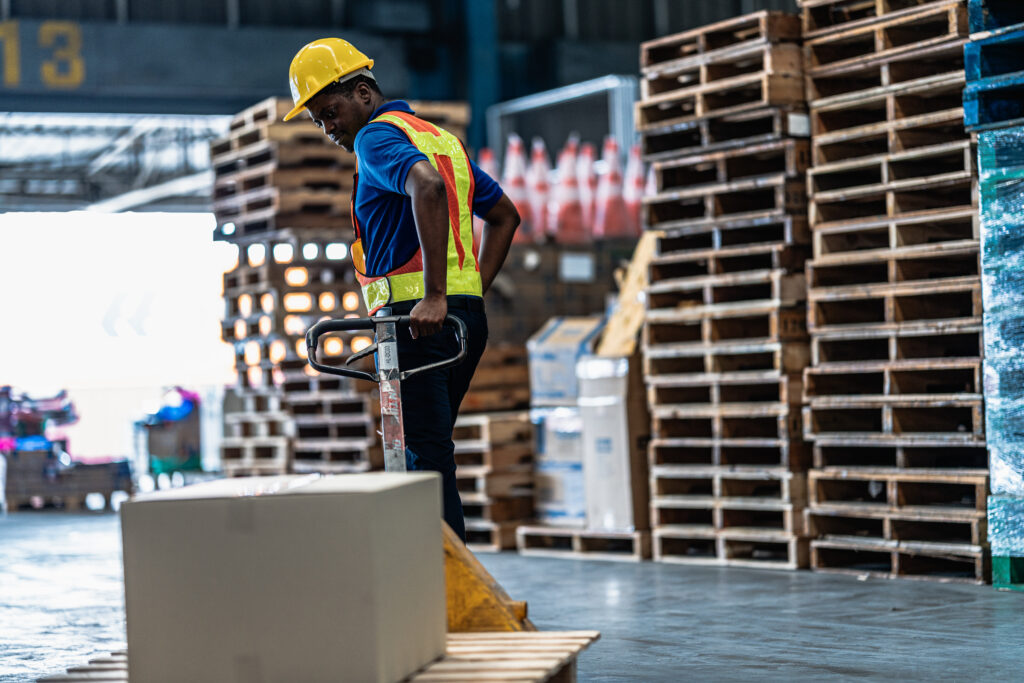A logistics worker pulling a hand pallet jack loaded with a cardboard box towards a shipping dock container inside a commercial storage warehouse.